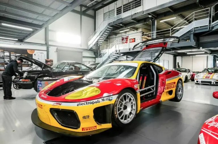 Voiture de course rouge et jaune avec portes papillon ouvertes, stationnée dans un garage moderne avec d’autres véhicules d’exception.